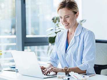 A female medical professional working on her laptop and smiling in her medical office.